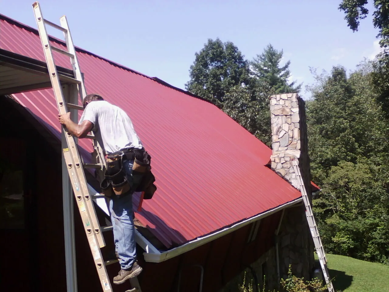 Roofer on a ladder beside a red metal roof and stone chimney — Soffit & Fascia Repair by Roofing Brothers in New Virginia